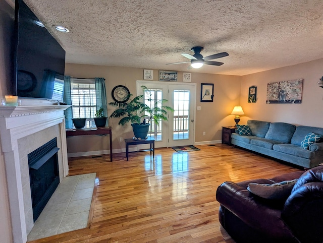 living room with a textured ceiling, ceiling fan, a tile fireplace, light wood finished floors, and baseboards