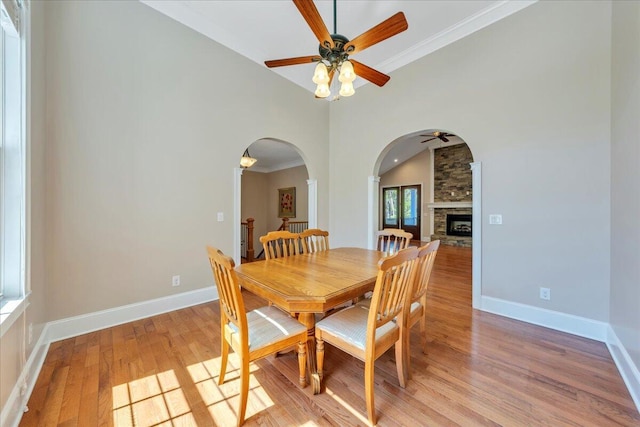 dining space with arched walkways, light wood-style floors, ceiling fan, a stone fireplace, and baseboards
