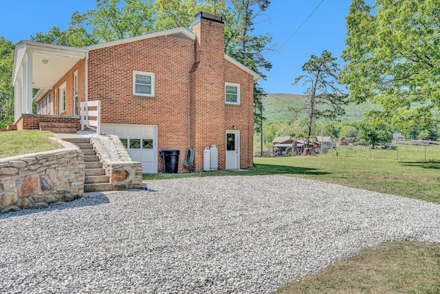 rear view of property featuring gravel driveway, a chimney, brick siding, an attached garage, and a yard