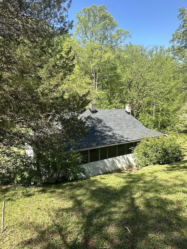 view of home's exterior featuring a wooded view, a chimney, roof with shingles, a yard, and a sunroom