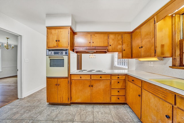 kitchen featuring brown cabinets, light countertops, a notable chandelier, under cabinet range hood, and white appliances