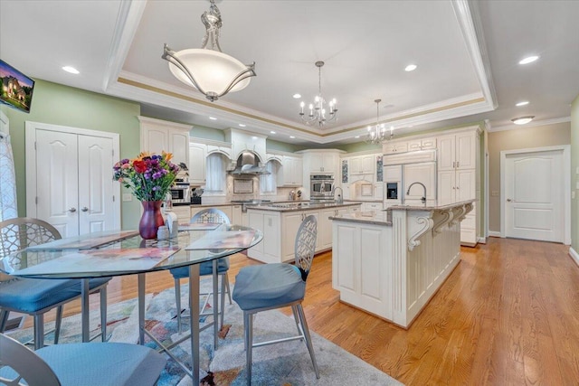 kitchen with a raised ceiling, a center island with sink, premium range hood, stainless steel appliances, and ornamental molding