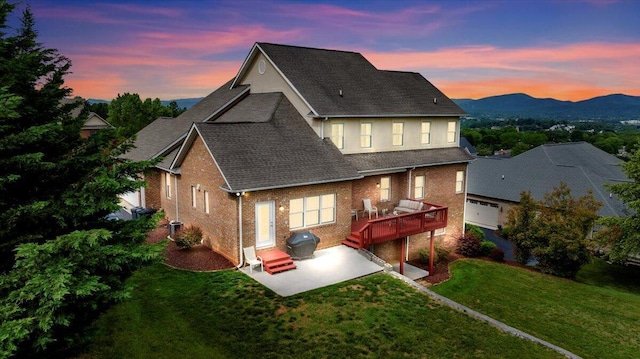 back of house at dusk with a patio, a yard, brick siding, a shingled roof, and a deck with mountain view