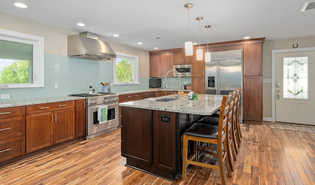 kitchen with wall chimney range hood, built in appliances, decorative backsplash, and light wood-style flooring