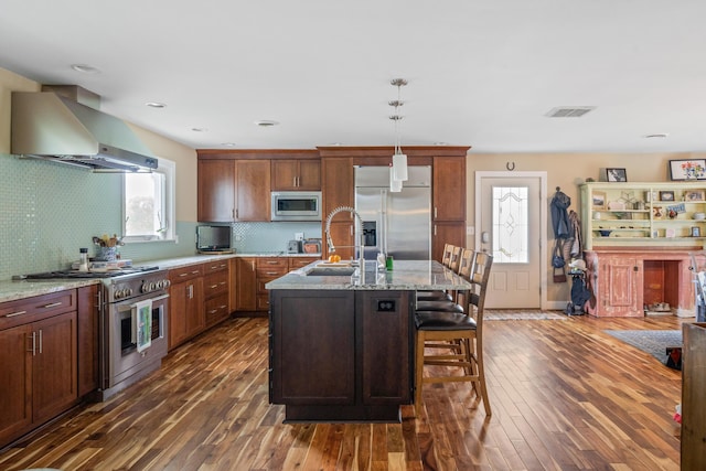 kitchen with brown cabinetry, backsplash, wood finished floors, light stone counters, and built in appliances