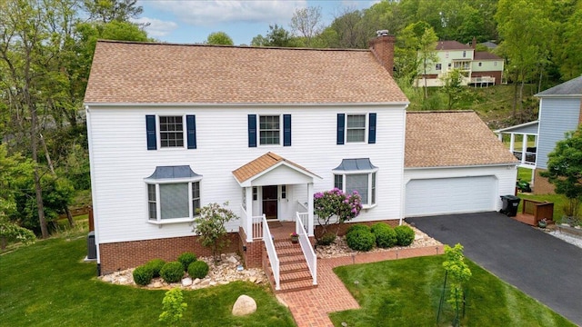 colonial inspired home featuring a shingled roof, aphalt driveway, a garage, a front yard, and a chimney