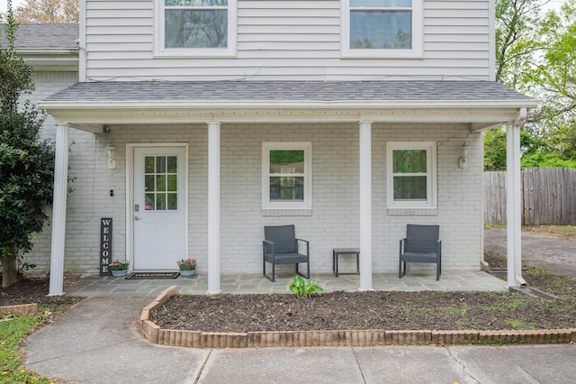view of exterior entry featuring a shingled roof, fence, brick siding, and covered porch