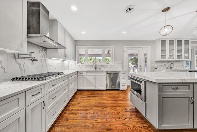 kitchen featuring wall chimney exhaust hood, appliances with stainless steel finishes, a sink, glass insert cabinets, and tasteful backsplash