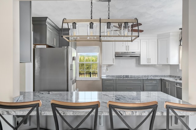 kitchen featuring a kitchen breakfast bar, stainless steel refrigerator with ice dispenser, backsplash, under cabinet range hood, and gray cabinetry