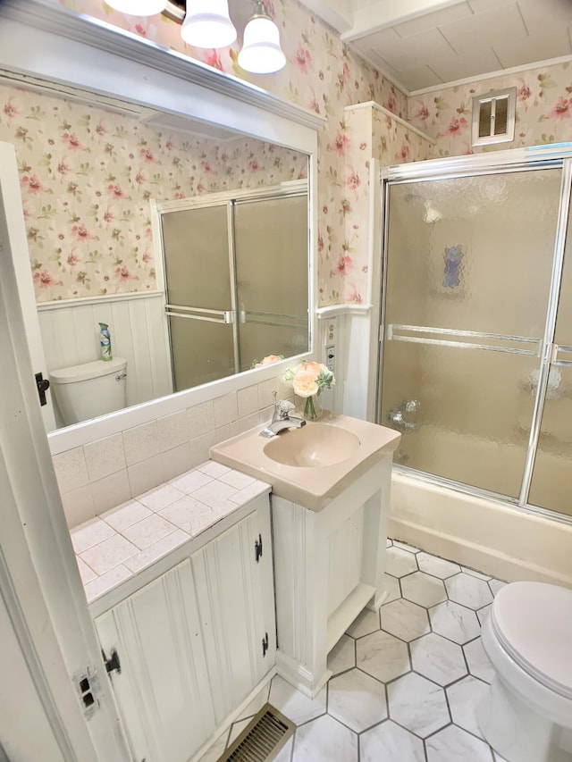 bathroom with wallpapered walls, toilet, vanity, a wainscoted wall, and combined bath / shower with glass door