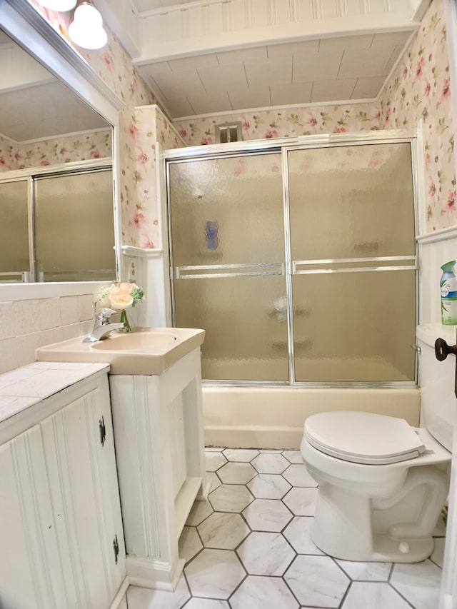 full bathroom with wallpapered walls, toilet, vanity, a wainscoted wall, and combined bath / shower with glass door