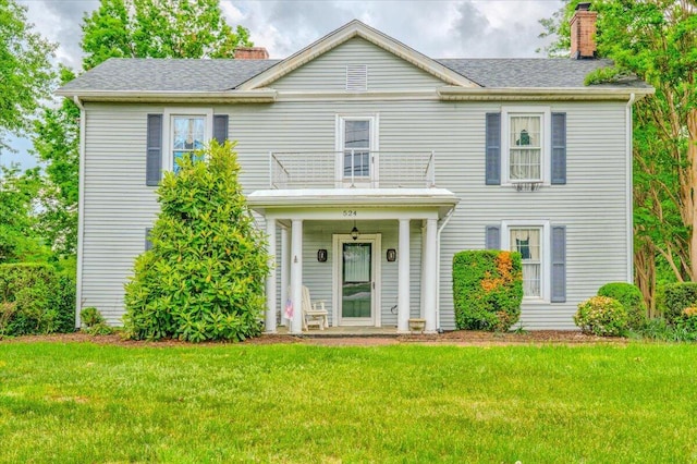 view of front of house featuring a shingled roof, a front lawn, a chimney, and a balcony