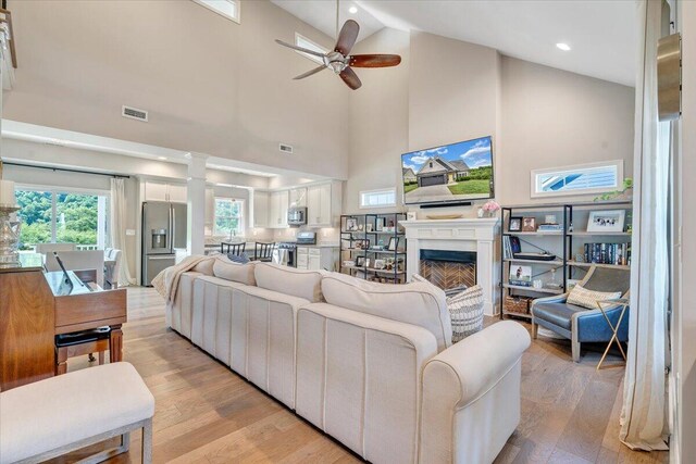 living room featuring a ceiling fan, a fireplace, light wood finished floors, high vaulted ceiling, and ornate columns