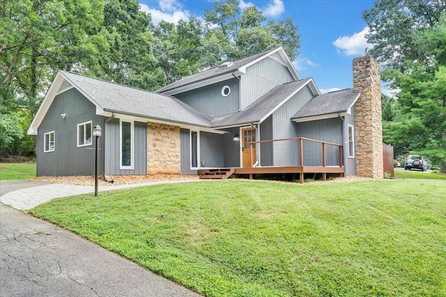 view of front of home with a front yard, a shingled roof, a chimney, stone siding, and a wooden deck