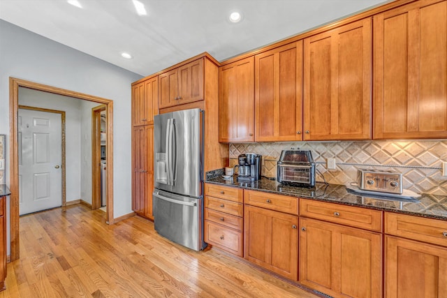 kitchen with stainless steel refrigerator with ice dispenser, brown cabinetry, dark stone counters, light wood finished floors, and backsplash