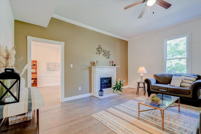 living area featuring light wood finished floors, baseboards, a ceiling fan, crown molding, and a glass covered fireplace