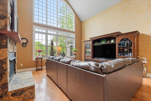 living room with high vaulted ceiling, light wood-type flooring, a stone fireplace, healthy amount of natural light, and baseboards