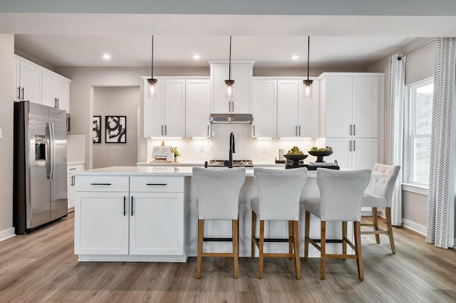 kitchen with stainless steel fridge, a center island with sink, under cabinet range hood, light countertops, and backsplash