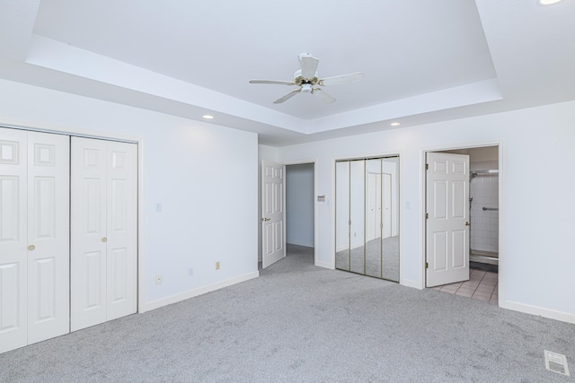 unfurnished bedroom with a raised ceiling, connected bathroom, ceiling fan, two closets, and light colored carpet