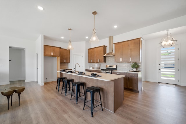 kitchen with light hardwood / wood-style floors, wall chimney range hood, stainless steel gas stove, hanging light fixtures, and a kitchen island with sink