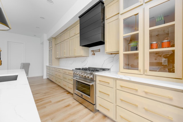 kitchen featuring stainless steel range, wall chimney exhaust hood, glass insert cabinets, light stone counters, and light wood-type flooring