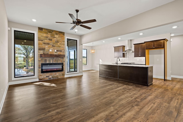 kitchen with a stone fireplace, a center island with sink, dark hardwood / wood-style floors, ceiling fan, and wall chimney range hood