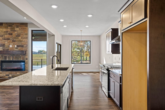 kitchen featuring stainless steel range with gas cooktop, light stone counters, a stone fireplace, and an island with sink