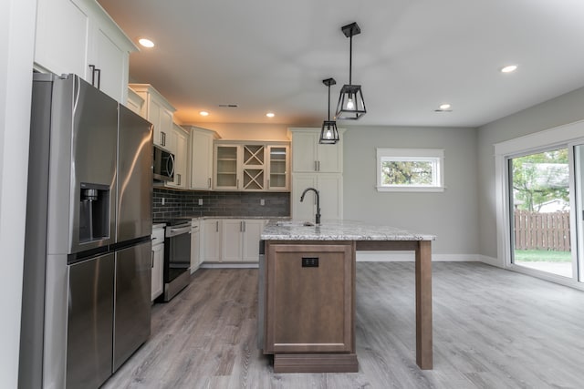 kitchen featuring a center island with sink, light stone counters, appliances with stainless steel finishes, decorative light fixtures, and light hardwood / wood-style flooring