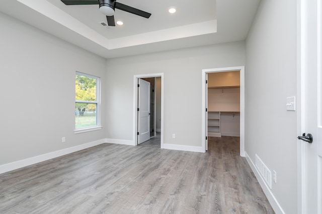 unfurnished bedroom with ceiling fan, a spacious closet, a raised ceiling, light hardwood / wood-style flooring, and a closet