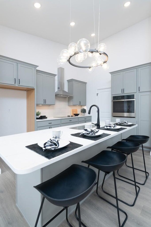 kitchen with hanging light fixtures, wall chimney range hood, an island with sink, oven, and light hardwood / wood-style floors