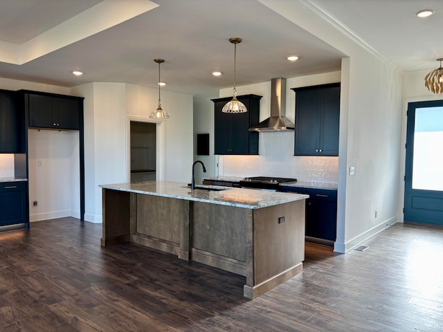 kitchen featuring a center island with sink, hanging light fixtures, a sink, light stone countertops, and wall chimney exhaust hood