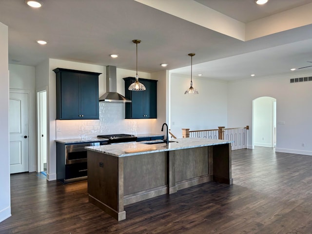 kitchen with arched walkways, a kitchen island with sink, a sink, wall chimney exhaust hood, and pendant lighting