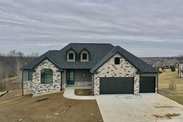 french country inspired facade with a garage, driveway, roof with shingles, and stone siding