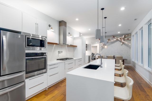 kitchen with stainless steel appliances, wall chimney range hood, sink, a kitchen island with sink, and light wood-type flooring