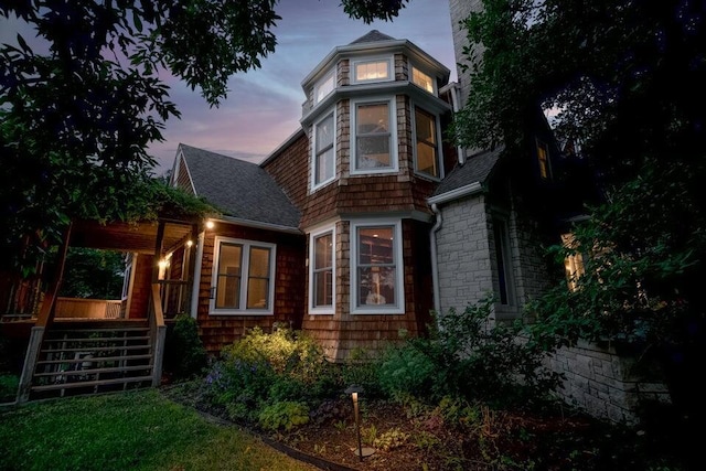 back of house at dusk with stone siding, a chimney, stairway, and roof with shingles