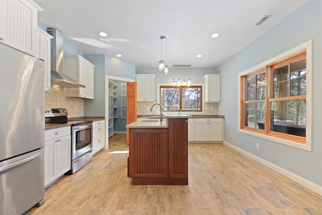 kitchen with stainless steel appliances, hanging light fixtures, white cabinets, wall chimney range hood, and an island with sink