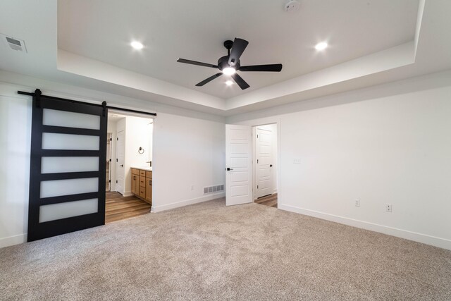 unfurnished bedroom featuring ceiling fan, ensuite bathroom, a tray ceiling, and a barn door