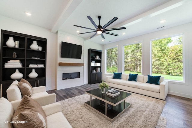 living room featuring ceiling fan, built in features, beam ceiling, dark wood-type flooring, and a fireplace