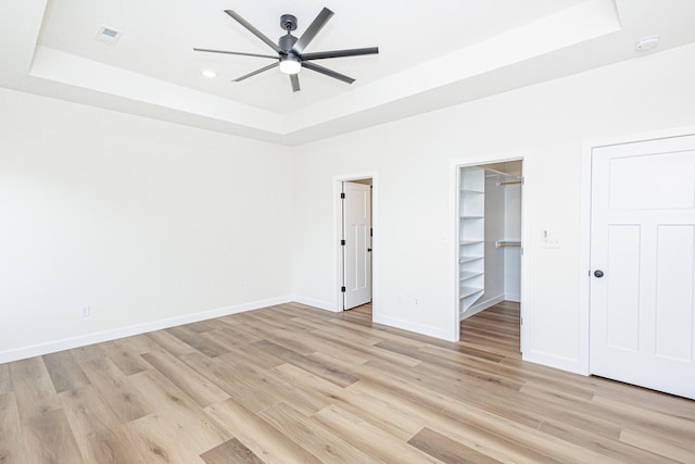 unfurnished bedroom featuring ceiling fan, light hardwood / wood-style flooring, a tray ceiling, and a walk in closet