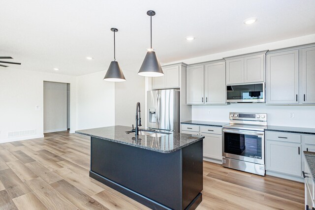 kitchen with dark stone counters, gray cabinets, a kitchen island with sink, appliances with stainless steel finishes, and light hardwood / wood-style floors