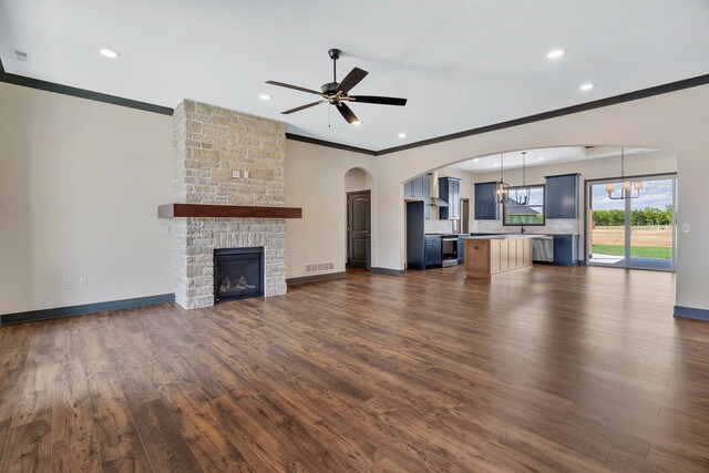 unfurnished living room with ceiling fan with notable chandelier, dark wood-type flooring, crown molding, and a stone fireplace