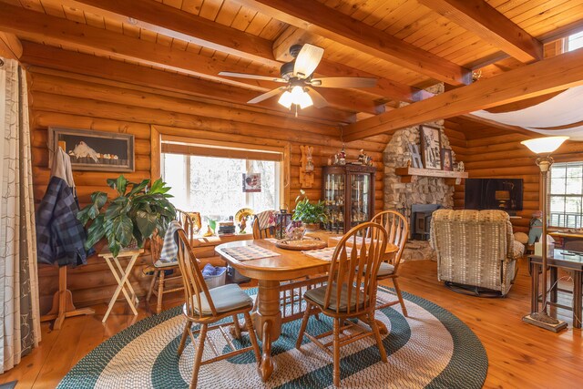 dining area featuring a stone fireplace, beam ceiling, light wood-type flooring, ceiling fan, and rustic walls