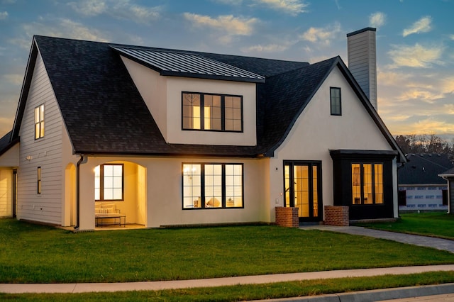 back of property at dusk featuring a chimney, stucco siding, roof with shingles, and a lawn