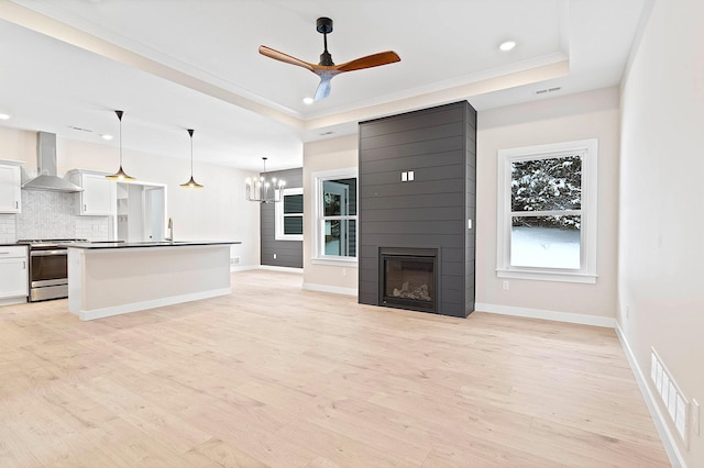 unfurnished living room featuring light wood-type flooring, a large fireplace, ceiling fan with notable chandelier, a raised ceiling, and sink