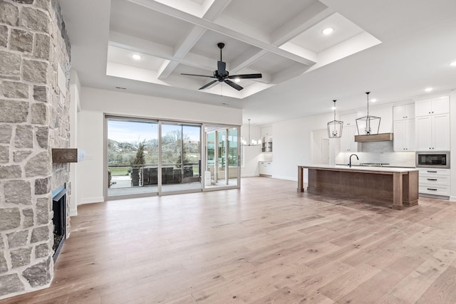 unfurnished living room featuring light wood-type flooring, beamed ceiling, ceiling fan with notable chandelier, coffered ceiling, and a stone fireplace