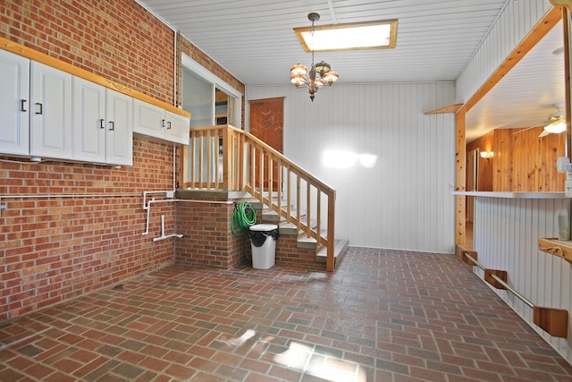 interior space featuring brick wall, ceiling fan with notable chandelier, pendant lighting, white cabinetry, and wood walls