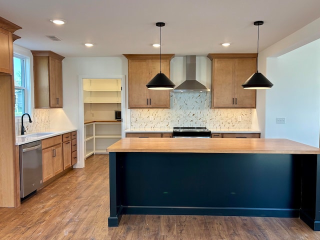 kitchen with wood counters, appliances with stainless steel finishes, hanging light fixtures, wall chimney range hood, and a sink