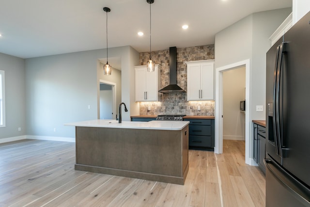 kitchen featuring black fridge with ice dispenser, a kitchen island with sink, sink, wall chimney range hood, and white cabinets