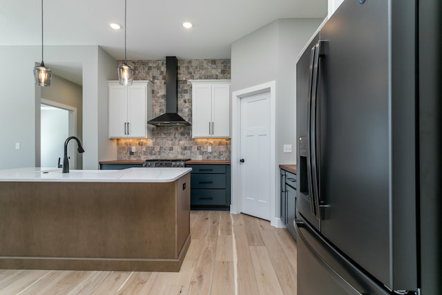 kitchen featuring black refrigerator with ice dispenser, wall chimney range hood, an island with sink, decorative light fixtures, and white cabinetry