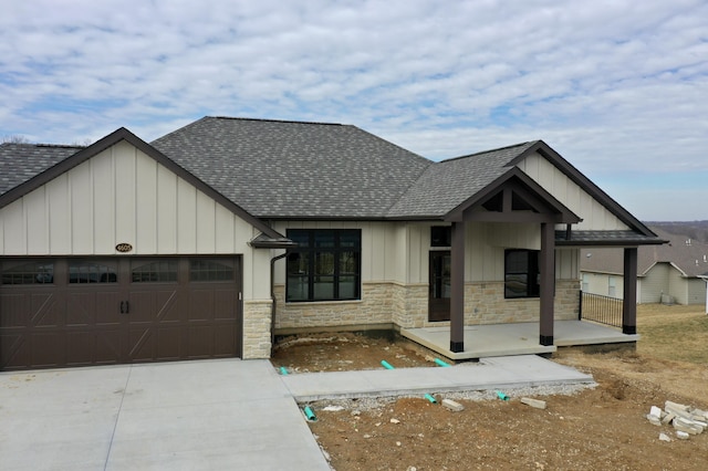 view of front of house with a shingled roof, stone siding, an attached garage, covered porch, and board and batten siding